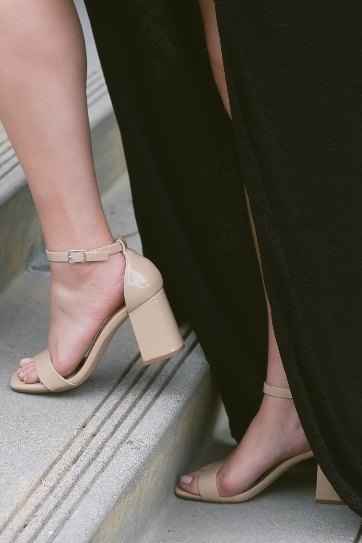 A beige block heel in a patent material with adjustable ankle strap worn by a woman in a black dress