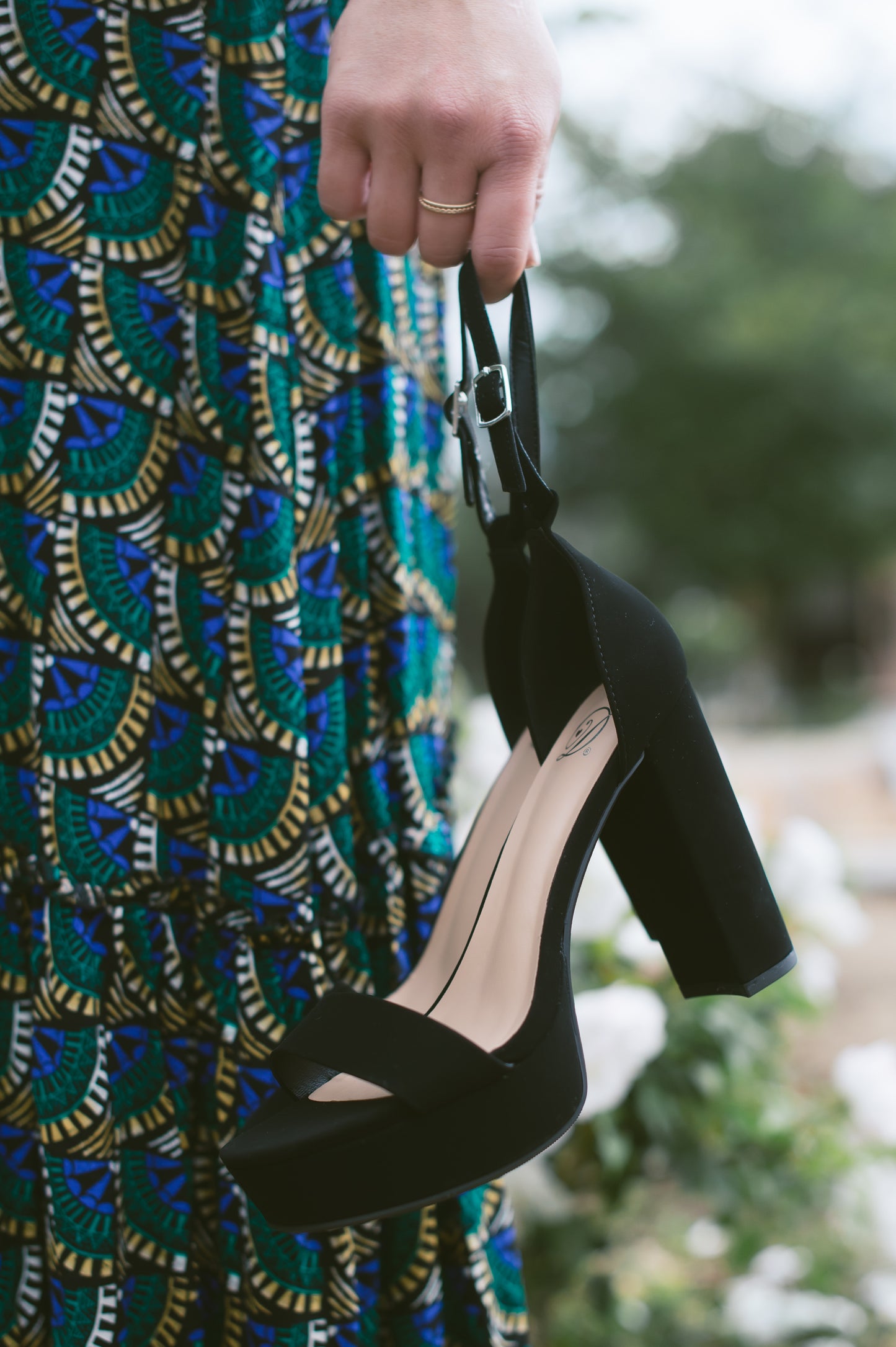 A black open-toe high block heel with a platform in a soft nubuck material, with an adjustable ankle strap and single strap across the toes held by a woman in a multicolor dress