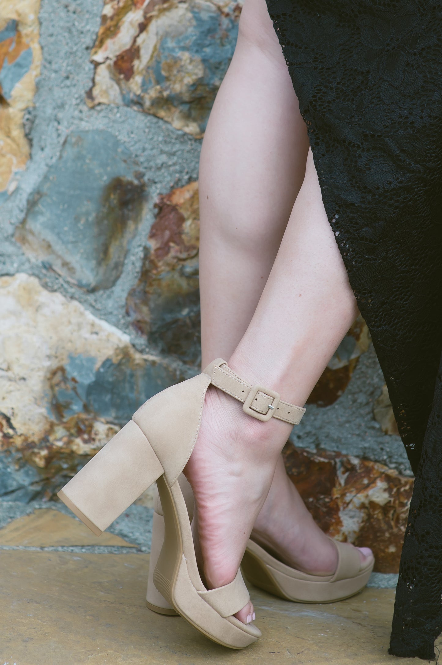 An open-toe block heel platform shoe in a natural tan color in soft nubuck material with a thick buckle on an adjustable ankle strap and single band across the toes worn by a woman in a black dress