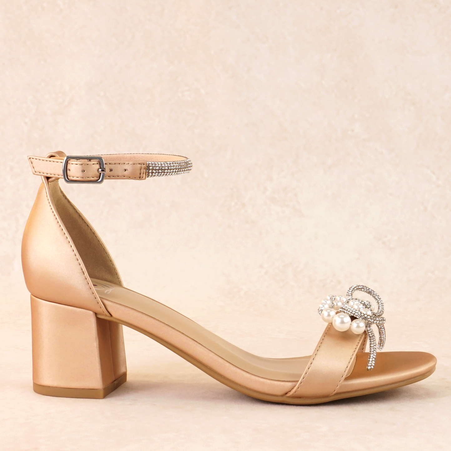 A champagne satin block heel with a pearl and rhinestone bow on a single strap across the toes and a rhinestone adjustable ankle strap