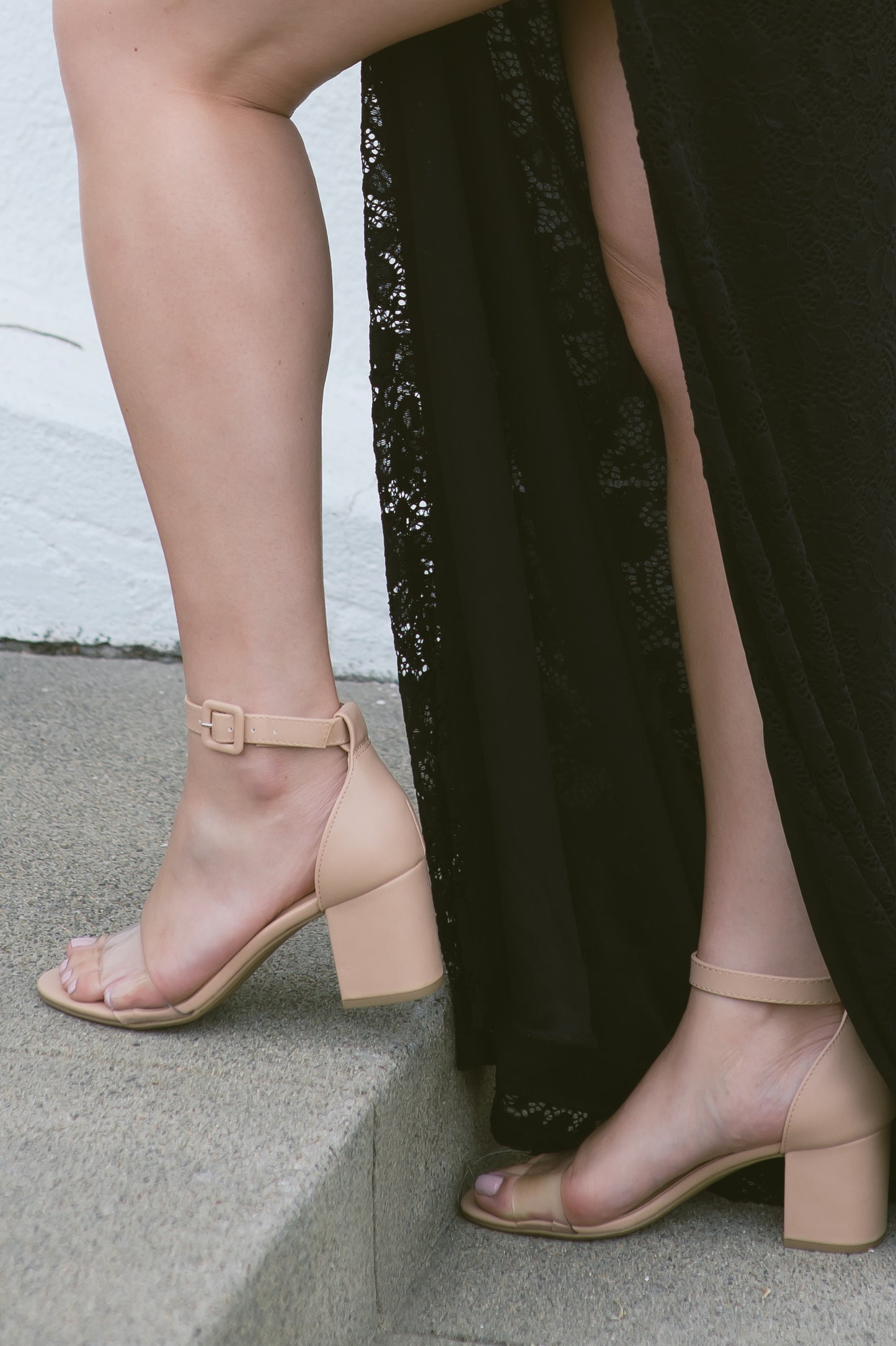 An open-toe block heel shoe in a natural tan color with a thick buckle on an adjustable ankle strap and single clear lucite band across the toes worn by a woman in a black dress