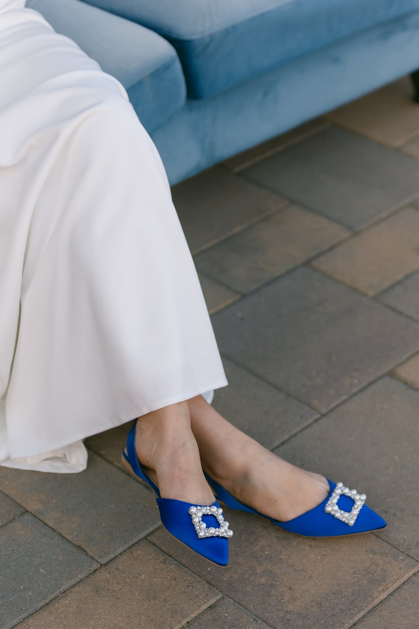 Slingback flat with a pearl buckle accent in a cobalt satin PU worn by a woman in a white dress