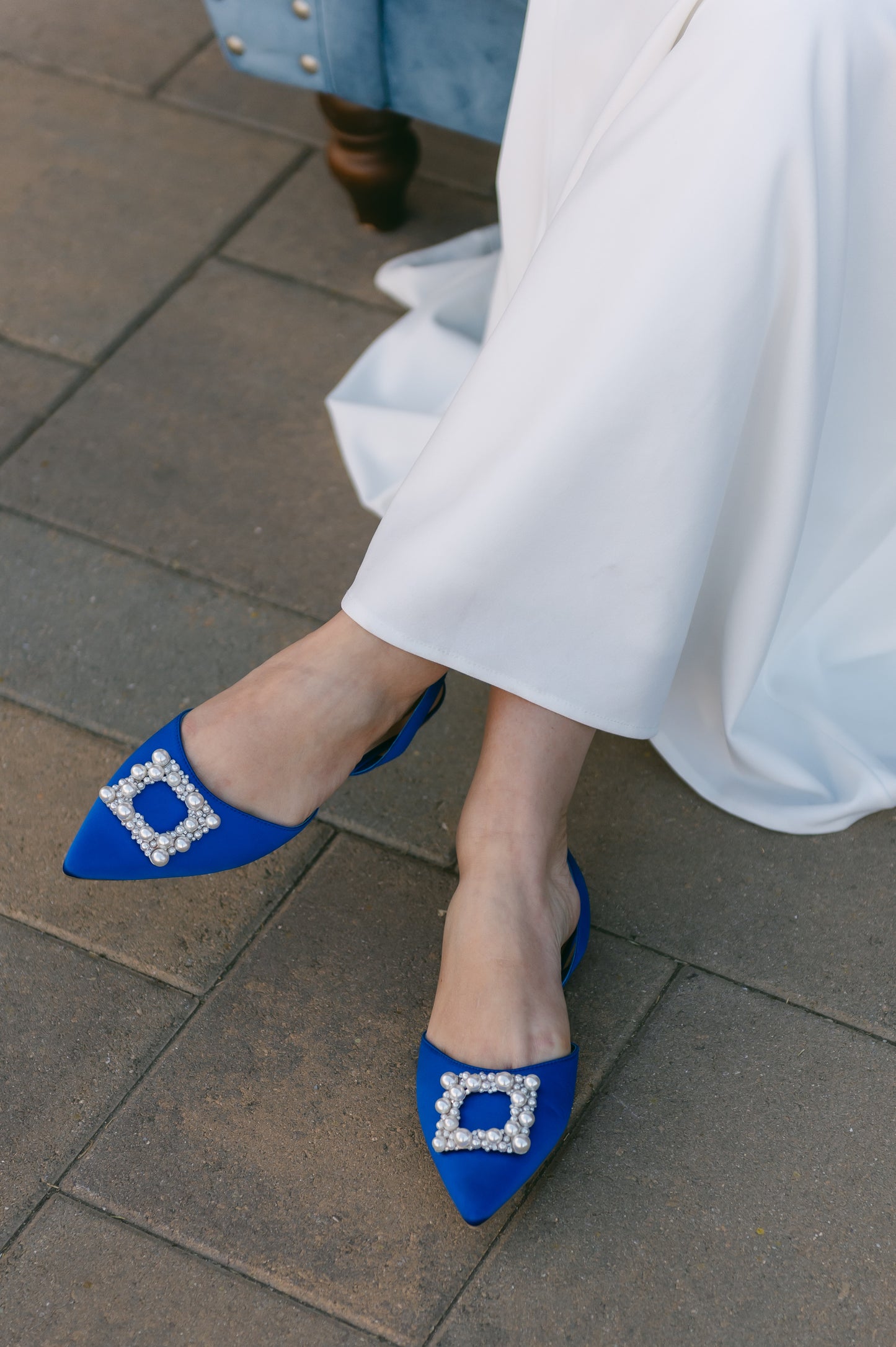 Slingback flat with a pearl buckle accent in a cobalt satin PU worn by a woman in a white dress