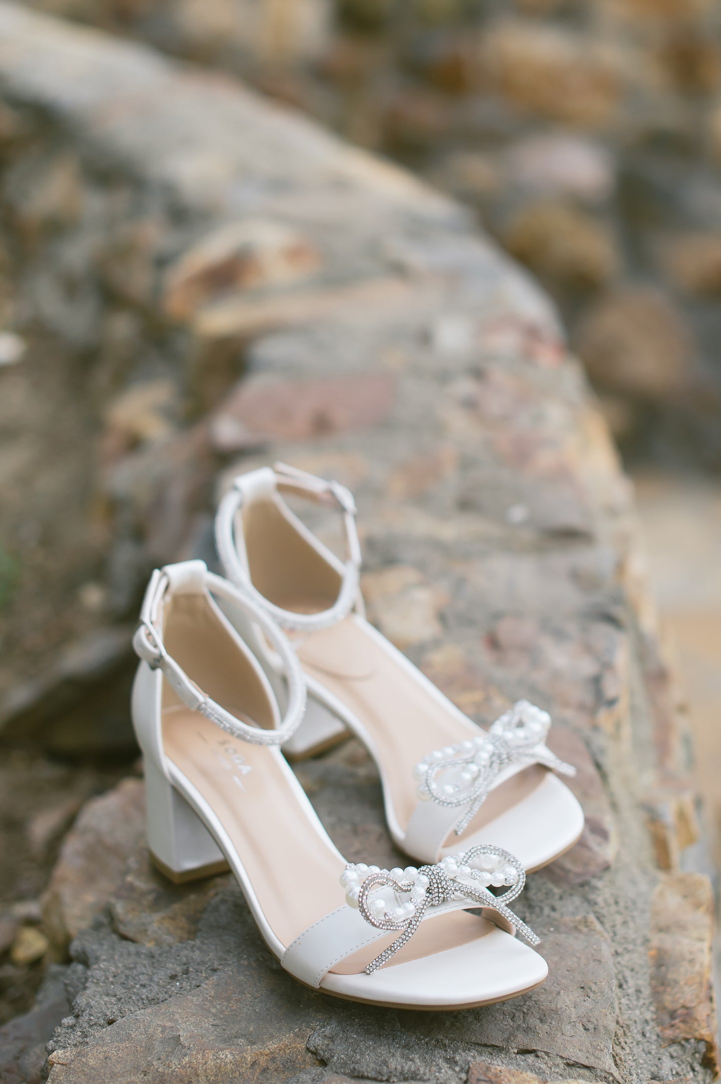 A white pearl block heel with a pearl and rhinestone bow on a single strap across the toes and a rhinestone adjustable ankle strap posed on brick