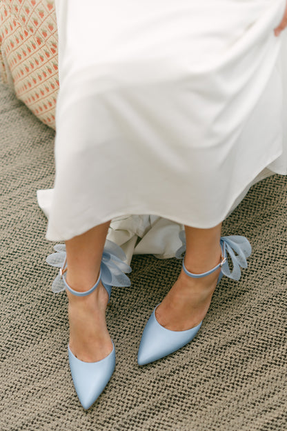 Blue d’orsay pump with ankle strap bow detail and a 3.25" stiletto heel worn by a woman in a white dress