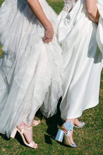 Flared 3" block heels in blue and pink with slim ankle strap and flower detail worn by two women in white dresses