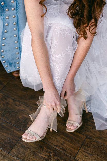 4" stiletto heel in gold with rhinestone band and wire mesh statement bow worn by a woman in a white dress