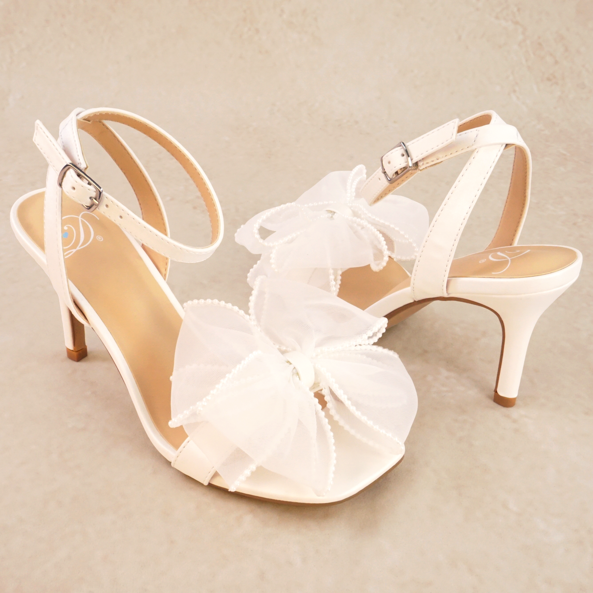 A pair of white pearlescent stiletto heels with a pearl studded mesh bow and adjustable ankle strap
