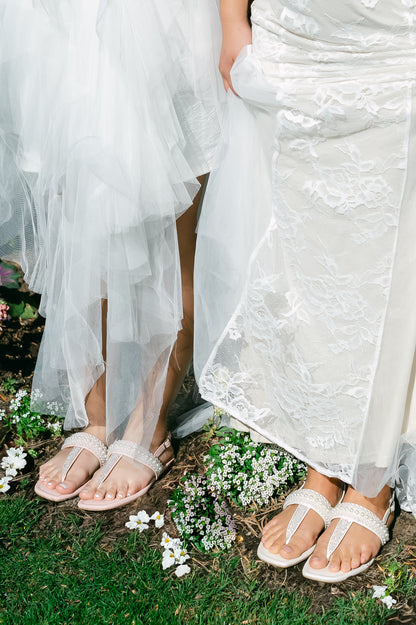 T-strap white and pink sandals with adjustable slingback and pearl and rhinestone accents being worn by two women in white dresses