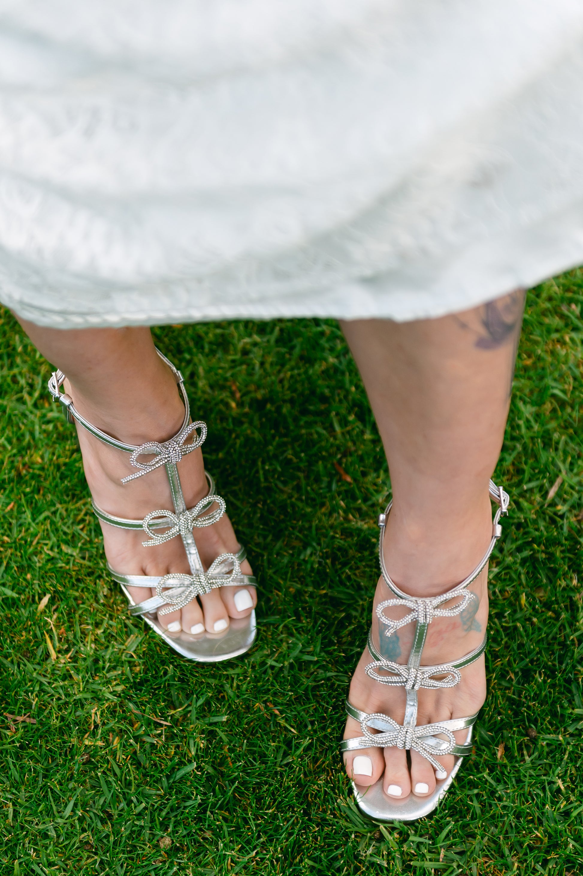 Rhinestone bows on a 4.25" block heel in metallic silver worn by a woman wearing a white dress