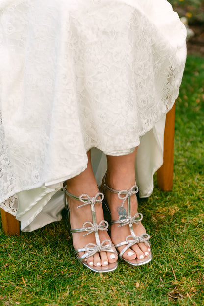 Rhinestone bows on a 4.25" block heel in metallic silver worn by a woman wearing a white dress