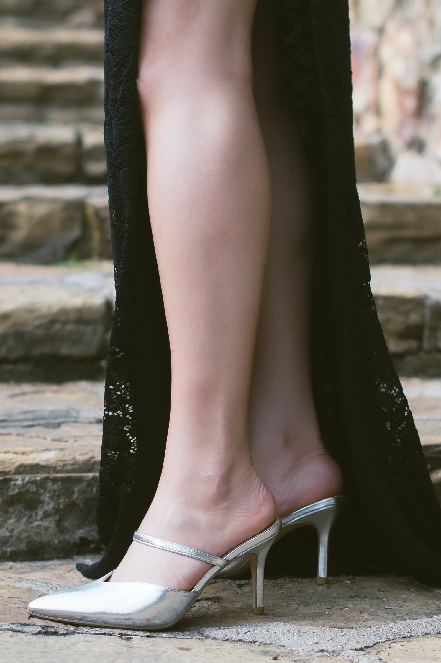 A silver closed-toe stiletto heel shoe in a shiny metallic material and an elasticized strap across the foot worn by a woman in a black dress