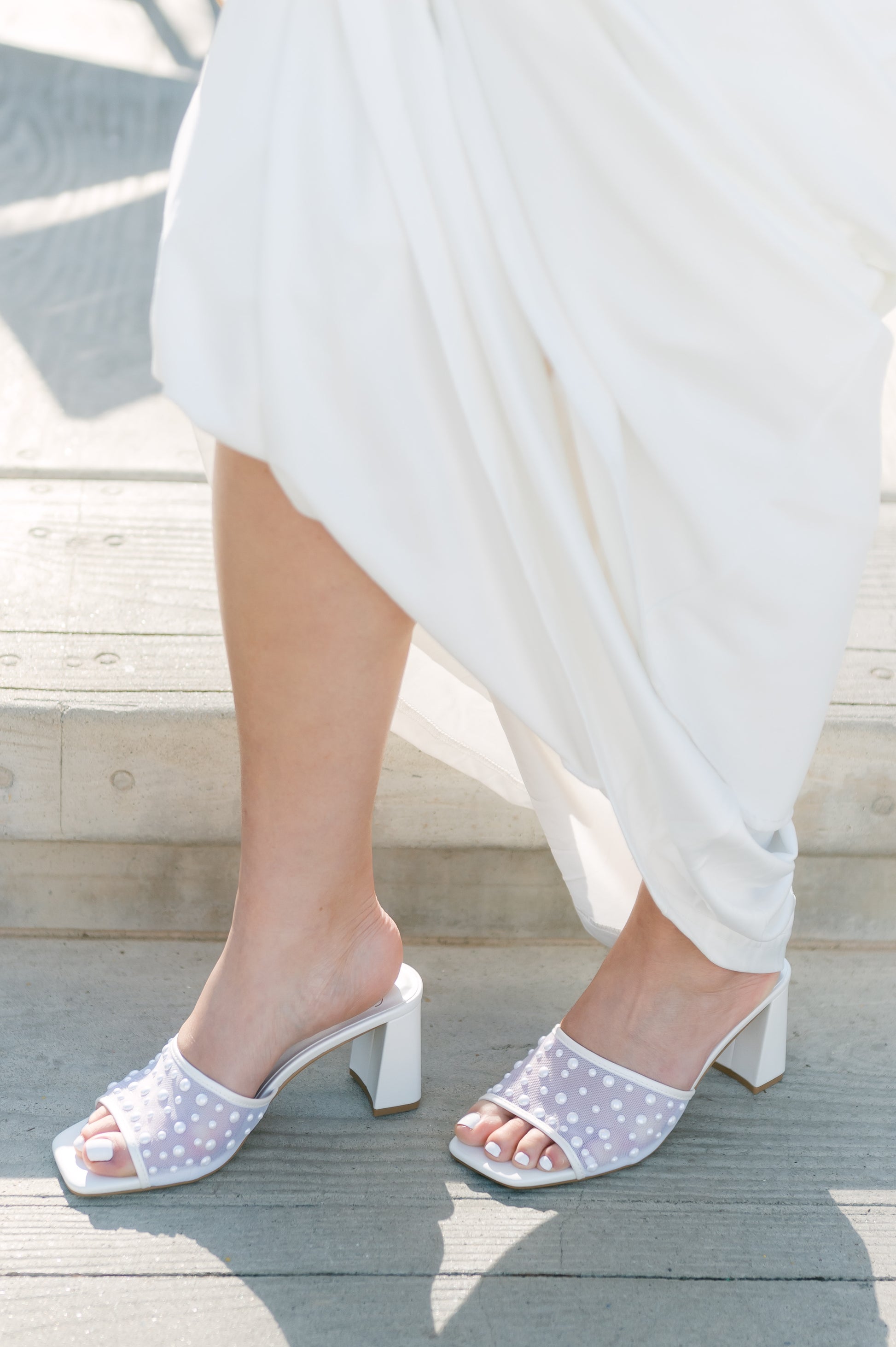 Mesh heeled white slide sandal with a 3" heel and pearls studded all over worn by a woman in a white dress