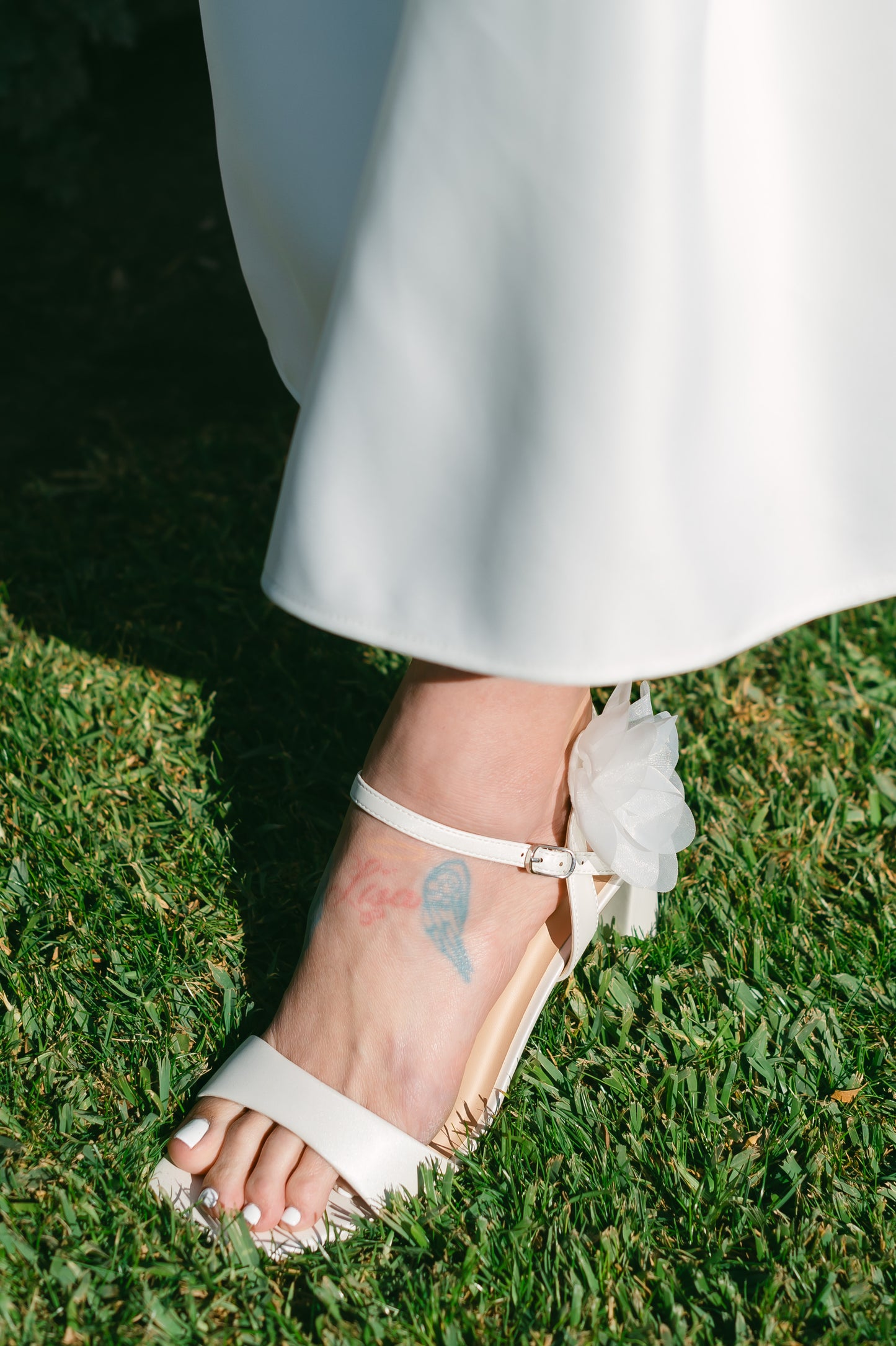 Flared 3" block heel in white with slim ankle strap and flower detail worn by a woman in a white dress