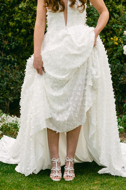 Pearl and rhinestone bows adorn a 4.25" block heel in pearlescent white worn by a woman in a white dress