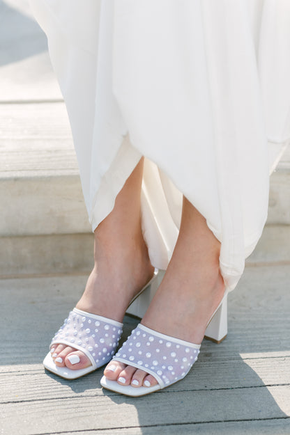 Mesh heeled white slide sandal with a 3" heel and pearls studded all over worn by a woman in a white dress