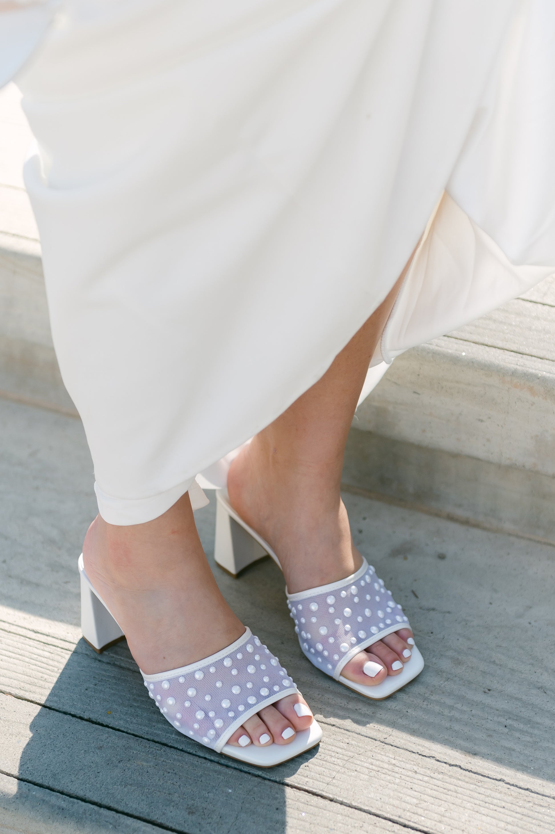 Mesh heeled white slide sandal with a 3" heel and pearls studded all over worn by a woman in a white dress