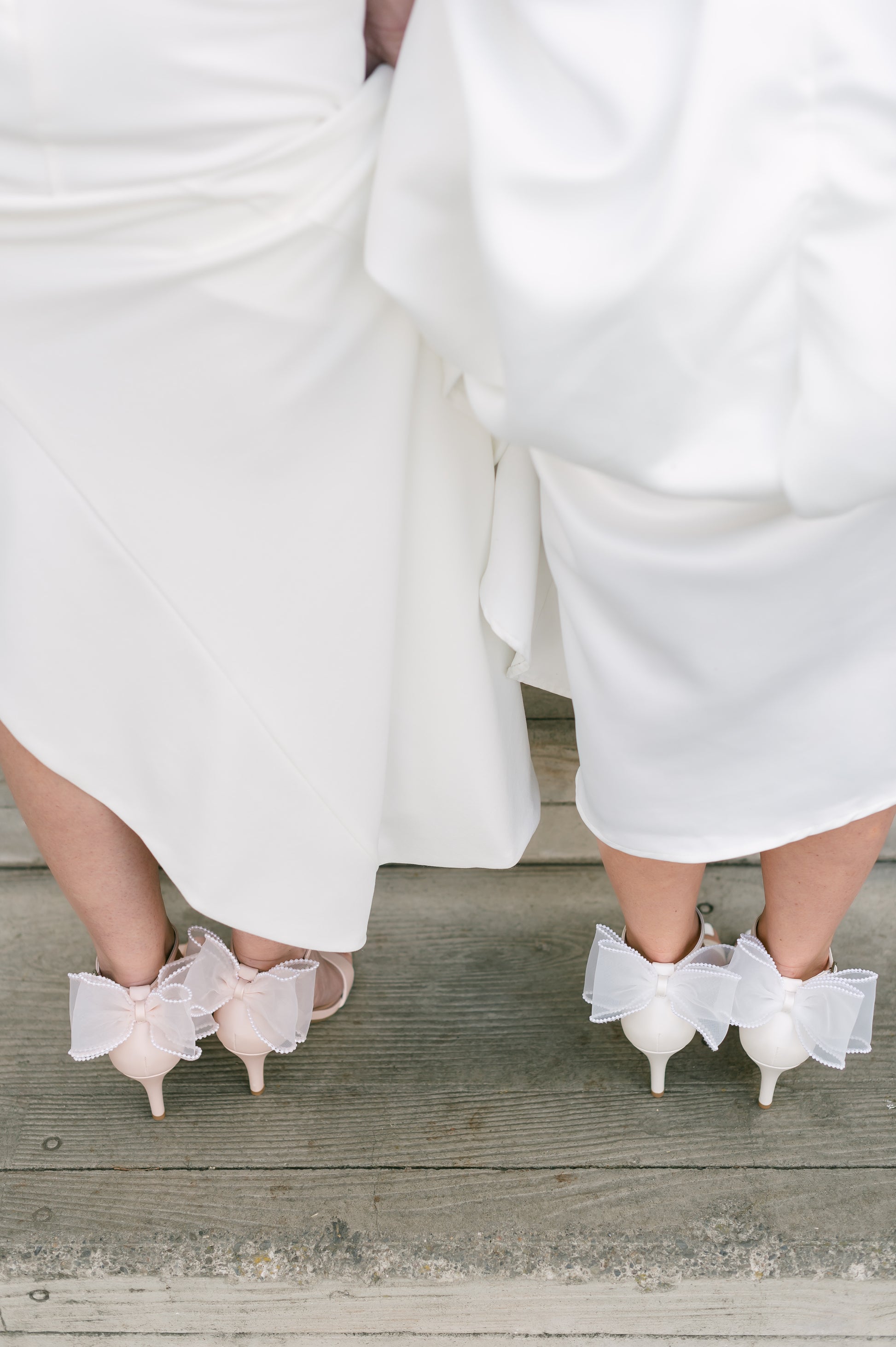 Simple single-band 3" stiletto heels in white and pink with a beaded bow on the back worn by two women in white dresses
