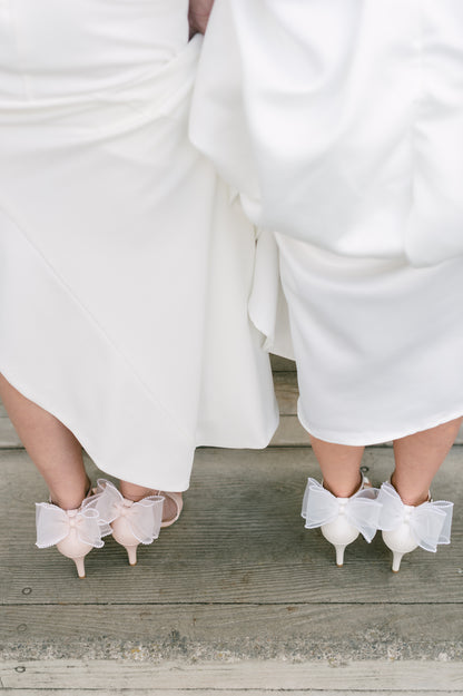 Simple single-band 3" stiletto heels in white and pink with a beaded bow on the back worn by two women in white dresses