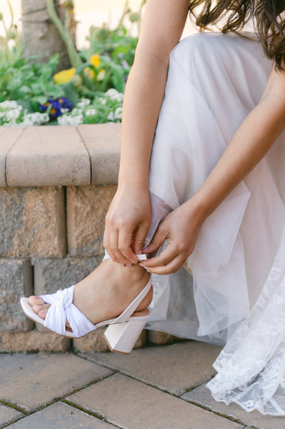 Pleated white satin knotted 3.25" block heel with ankle strap worn by a woman in a white dress